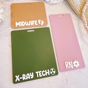 Peut inclure: Trois badges rectangulaires : un beige avec "MIDWIFE" et des empreintes de pieds, un vert avec "X-RAY TECH" et un symbole de radiation, et un rose avec "RN" et une fleur.