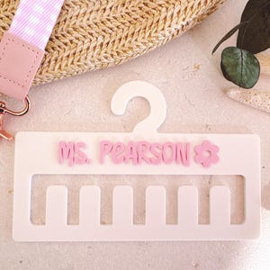 May include: A white acrylic lanyard holder with the name "Ms. Pearson" in pink letters and a pink flower. A pink and white gingham lanyard with a rose gold clasp is attached. The holder has slots for multiple lanyards.