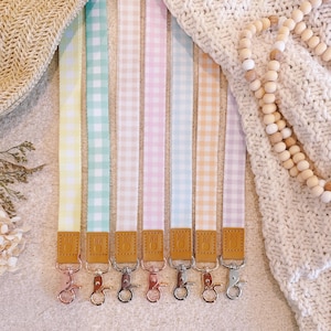 May include: Seven fabric lanyards featuring a gingham pattern in pastel yellow, mint green, cream, pink, light blue, peach, and lavender. Each strap includes a tan leather tab with a metal lobster clasp in rose gold or silver finish.