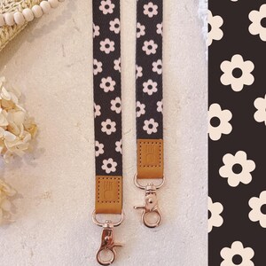 May include: Two black and white daisy patterned lanyards with rose gold swivel clasps and brown leather accents. The lanyards are hanging vertically against a light background. The daisy pattern is also visible on the right side of the image.