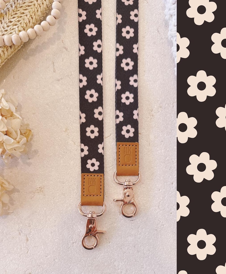 May include: Two black and white daisy patterned lanyards with rose gold swivel clasps and brown leather accents. The lanyards are hanging vertically against a light background. The daisy pattern is also visible on the right side of the image.