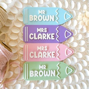 Op de afbeelding: Vier kleurrijke, potloodvormige naamstickers met witte letters. De stickers lezen "Mr. Brown", "Mrs. Clarke", "Mrs. Clarke" en "Mr. Brown".