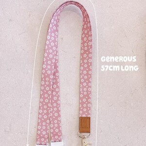 May include: Pink floral lanyard with a metal clasp and leather tag. The lanyard is 57cm long. The lanyard features a repeating pattern of small white flowers on a pink background. The text "Generous" is also visible.