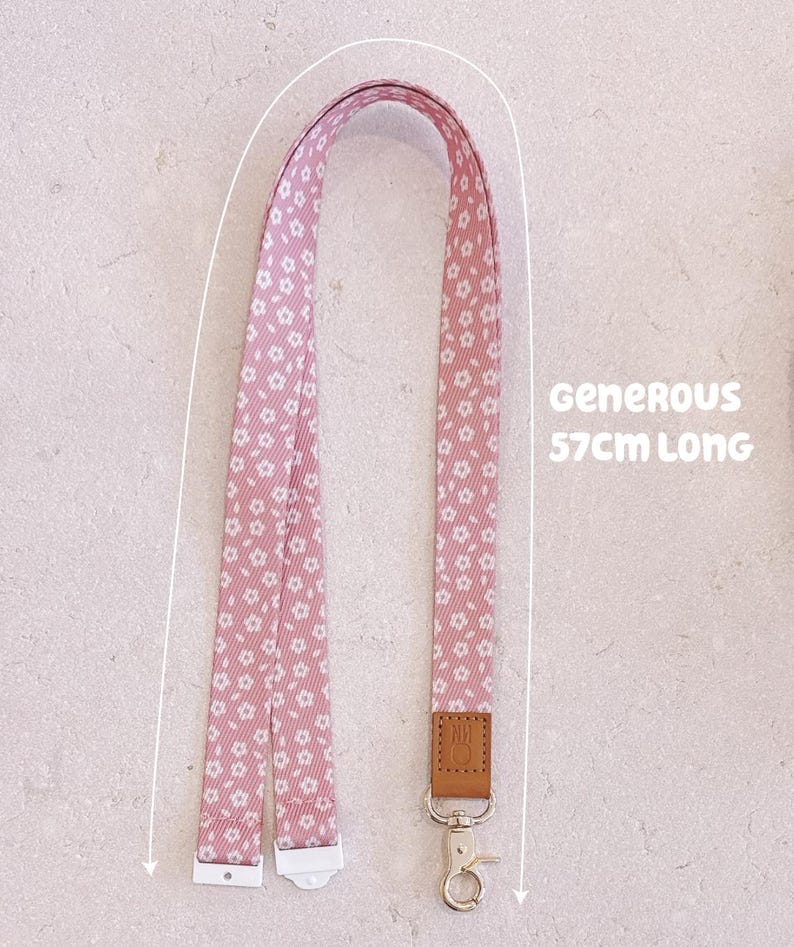 May include: Pink floral lanyard with a metal clasp and leather tag. The lanyard is 57cm long. The lanyard features a repeating pattern of small white flowers on a pink background. The text "Generous" is also visible.