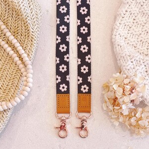 May include: Two black and white daisy patterned lanyards with rose gold hardware. The lanyards have brown leather accents and are displayed on a neutral background with a knitted hat and beaded necklace.