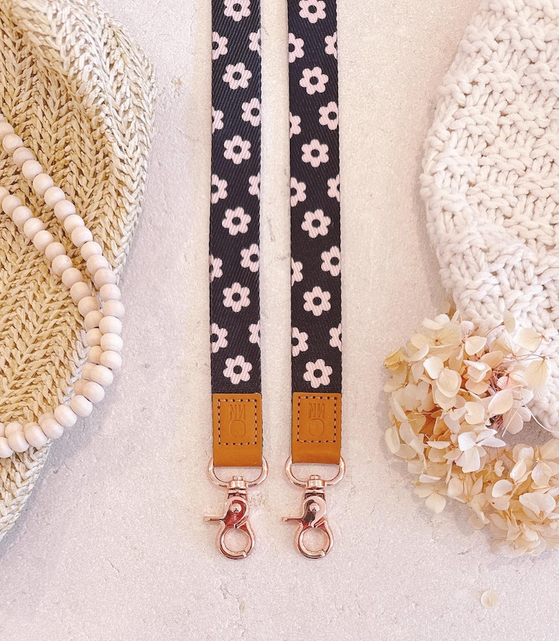 May include: Two black and white daisy patterned lanyards with rose gold hardware. The lanyards have brown leather accents and are displayed on a neutral background with a knitted hat and beaded necklace.