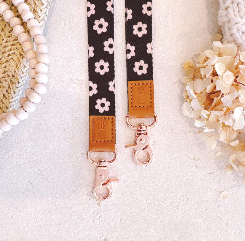 May include: Two black and white floral patterned straps with rose gold clasps. The straps have brown leather accents with the initials "NM" embossed. The background is a soft white with decorative elements.