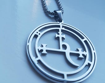 Puede incluir: Collar con colgante plateado con un diseño circular con un símbolo estilizado. El colgante cuelga de una cadena a juego. El diseño incluye formas de cruz y líneas curvas dentro del círculo. El collar se muestra sobre un fondo azul claro.