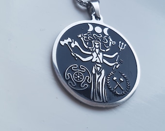 Peut inclure: Un collier pendentif argenté avec un pendentif circulaire. Le pendentif a un fond noir et représente une figure à plusieurs bras avec des croissants de lune au-dessus. Divers symboles entourent la figure, dont un marteau, un trident et une couronne.