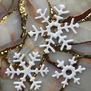May include: Three white ceramic snowflake ornaments with silver string hangers. The snowflakes are on a background of gold and white marble.