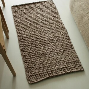 Könnte beinhalten: Ein rechteckiger, handgestrickter Teppich in einem neutralen Braunton. Der Teppich hat eine strukturierte Oberfläche und ein schlichtes, minimalistisches Design. Er liegt auf einem hellen Boden, mit einem Teil eines Holzstuhls und einem beigen Kissen.