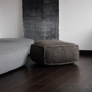 May include: A dark gray knitted ottoman with a square shape sits on a dark wood floor. A gray sofa is in the background.