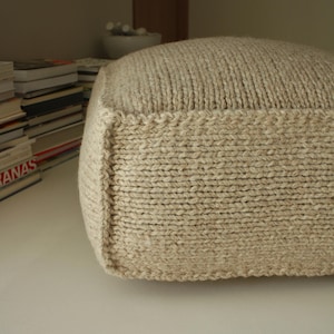 May include: A square, beige knitted pouf. The pouf is made of thick yarn and has a textured surface. The edges are neatly finished with a decorative stitch. The pouf is a neutral color and would complement any decor.