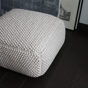 Op de afbeelding: Een vierkante, crèmekleurige gebreide poef met een getextureerd oppervlak. De poef staat op een donkerbruine houten vloer. Op de achtergrond is een ingelijste foto en een grijs geschilderde muur.