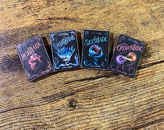 May include: Four miniature book replicas featuring black covers with colorful graphic illustrations and titles. The books are titled Lightlark, Nightbane, Skyshade, and Crowntide, all authored by Alex Aster, displayed in a horizontal row.