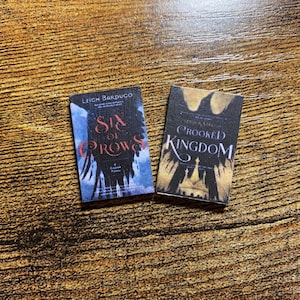May include: Two paperback books, Six of Crows and Crooked Kingdom, by Leigh Bardugo. The covers feature dark blue and black backgrounds with illustrations of crows and a crown.