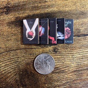 May include: A set of five paperback books with black covers and red accents. The titles are "Twilight", "New Moon", "Eclipse", "Dawn", and "Midnight Sun".
