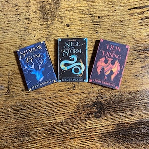 May include: Three miniature books with dark blue, black, and red covers. The books are titled "Shadow and Bone", "Siege and Storm", and "Ruin and Rising" by Leigh Bardugo. Each book features a different illustration: a deer, a snake, and a phoenix.