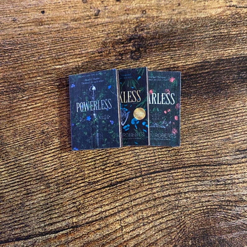 Pode incluir: Tr&ecirc;s livros de bolso com capas pretas e desenhos florais. Os t&iacute;tulos s&atilde;o "Powerless", "Kless" e "Arless" de Jennifer L. Armentrout.