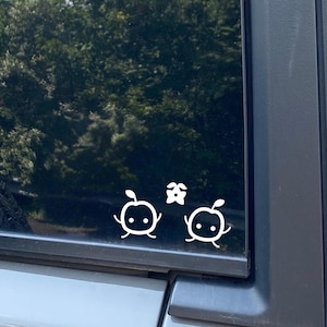 May include: A black car window with a white sticker of two cartoon characters with a flower between them.
