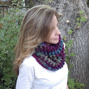 May include: A handmade crochet cowl in shades of purple, teal, and green. The cowl is worn around the neck and is made of a textured yarn. The background includes a tree trunk and green foliage.