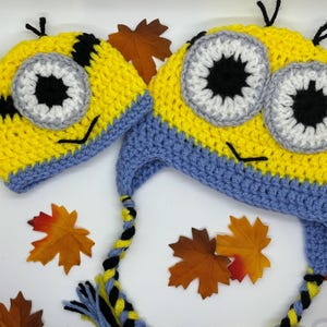 May include: Two crocheted Minion hats, one larger than the other, with yellow and blue yarn. The hats have black and white eyes and a black mouth.
