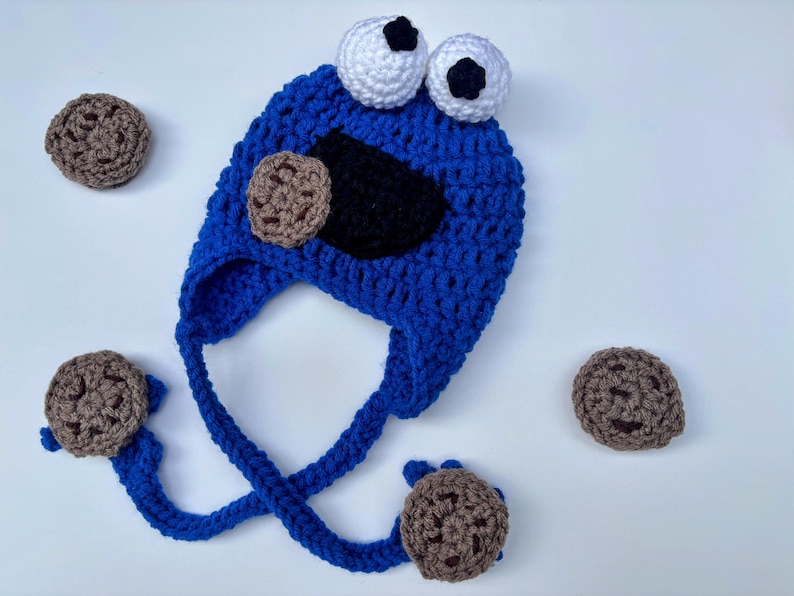 May include: A handmade blue crochet hat with white eyes and a black mouth, resembling a cartoon character. The hat has ear flaps with attached brown crochet cookies. Additional brown crochet cookies are scattered around the hat.