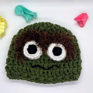 May include: A green crocheted hat with large white-rimmed eyes and a black smiling mouth. The hat has brown fuzzy accents above the eyes. Colorful crumpled paper is scattered around the hat.