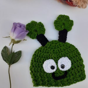 May include: Green crocheted hat with black antennas, white eyes, and a black smiling mouth. The hat is shaped like a cartoon alien.