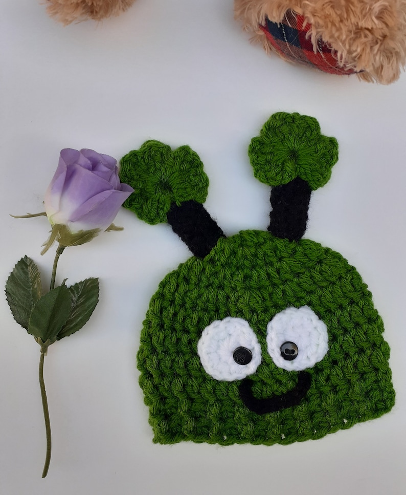 May include: Green crocheted hat with black antennas, white eyes, and a black smiling mouth. The hat is shaped like a cartoon alien.