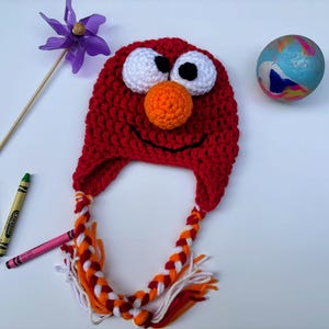 May include: A handmade red crochet hat with white eyes, black pupils, and an orange nose, resembling a cartoon character. The hat has braided tassels in orange, white, and red. Crayons and a pinwheel are also in the image.