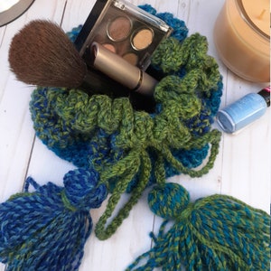 May include: A green and blue crocheted drawstring bag with two tassels. The bag is filled with makeup brushes and a compact eyeshadow palette.