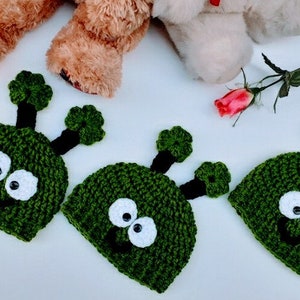 May include: Three green crocheted hats with white eyes and black smiles. The hats have two green antennas on top.