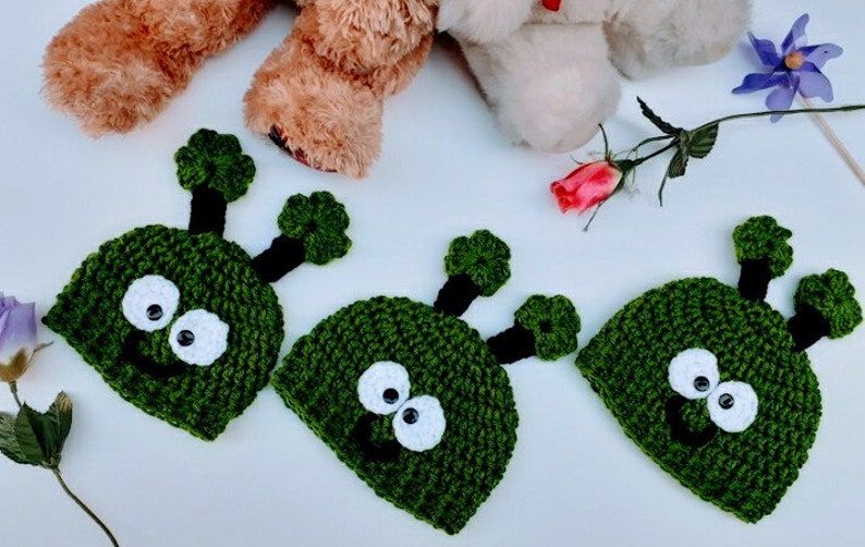 May include: Three green crocheted hats with white eyes and black smiles. The hats have two green antennas on top.