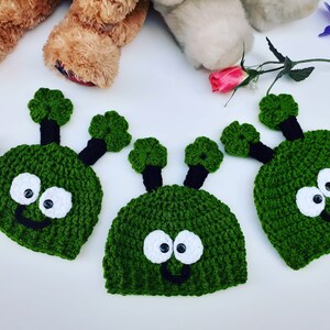 May include: Three green crocheted alien hats with black antennas and white eyes. The hats have a smiling mouth.