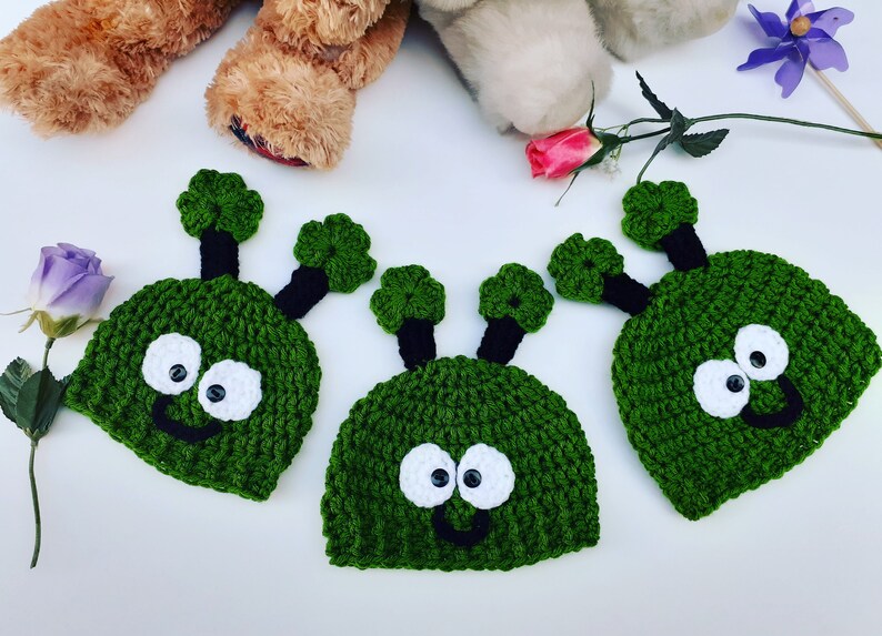 May include: Three green crocheted alien hats with black antennas and white eyes. The hats have a smiling mouth.