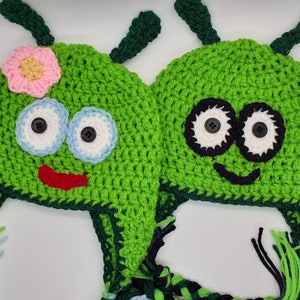 May include: Two green crocheted hats with whimsical faces. One hat has a pink flower and the other has a smiling face with black eyes. Both hats have green and black tassels.