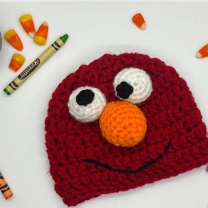 May include: A handmade red crochet hat with white and black eyes, and an orange nose. The hat has a black stitched smile. Crayola crayons and candy corn are scattered around the hat.