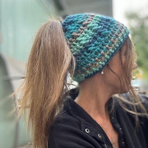 May include: A blue, green, and brown crocheted hat with a ponytail hole. The hat is made with a chunky yarn and has a loose, slouchy fit.