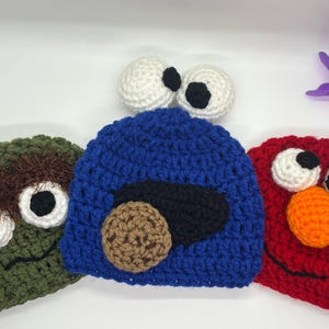 May include: Three handmade crochet hats in the style of popular characters. The hats are green, blue, and red, with white and black eyes, and other character-specific details. The hats are designed to resemble a popular children's show.