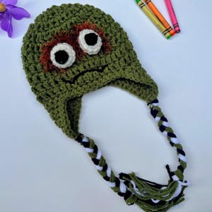 May include: A handmade green crochet hat with a monster face design. The hat features large white eyes with black pupils, orange fur accents, and a stitched mouth. Braided tassels in black, white, and green hang from the ear flaps. A purple pinwheel and crayons are in the background.