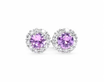May include: A pair of stud earrings featuring round, faceted light purple gemstones at the center. Each stone is surrounded by a halo of small, clear, sparkling white stones set in a polished silver-toned metal frame with a classic circular design.
