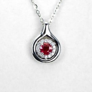 May include: A silver pendant necklace with a teardrop-shaped design. The pendant features a central, round, deep red gemstone, encircled by a halo of smaller, clear gemstones. The necklace chain is also silver.