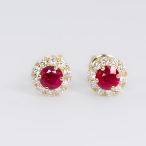 May include: A pair of gold earrings with a red ruby gemstone surrounded by a halo of smaller white gemstones.
