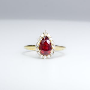 Puede incluir: Un anillo de rubí en forma de pera con un halo de pequeños diamantes engastados en oro amarillo.
