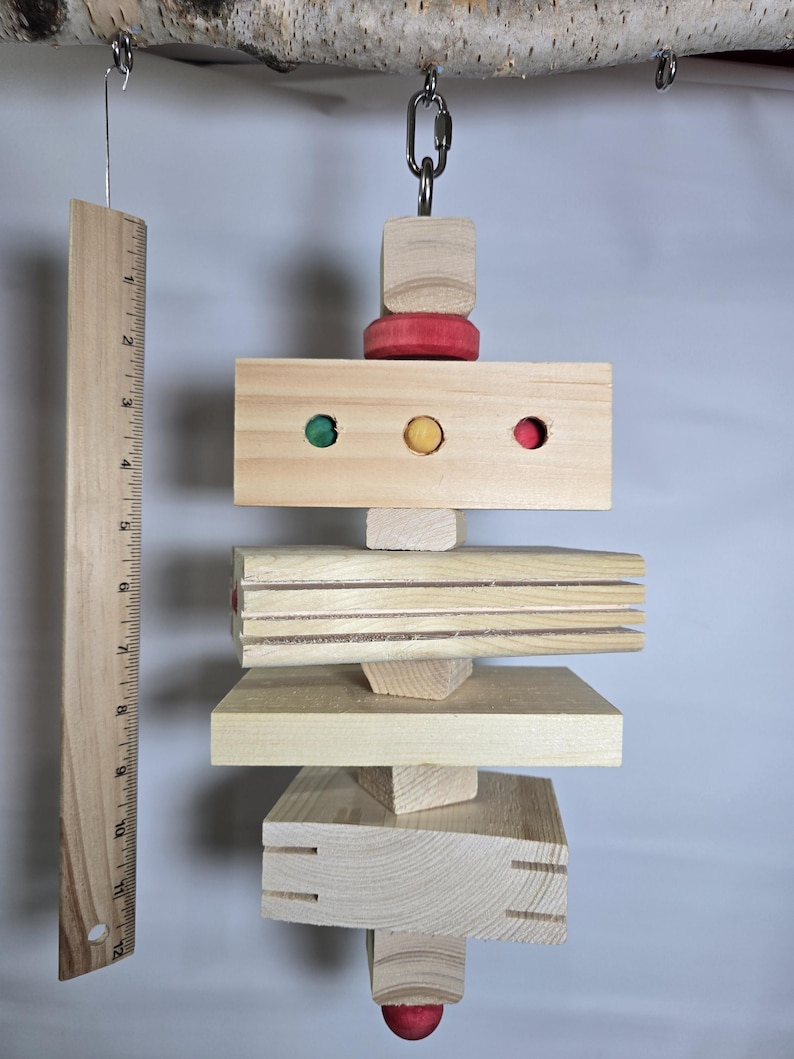 Puede incluir: Un juguete de madera para p&aacute;jaros con varias formas y colores. El juguete est&aacute; suspendido de una rama y presenta una regla para la comparaci&oacute;n de tama&ntilde;os. El juguete incluye detalles en rojo, verde y amarillo.