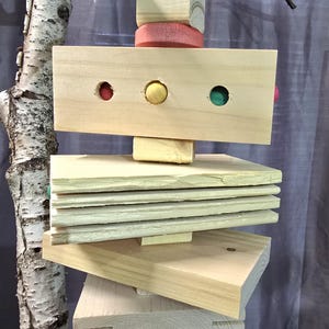 Puede incluir: Un juguete colgante para p&aacute;jaros hecho de madera natural, con una variedad de formas y colores. El juguete incluye un cubo de madera, un disco rosa y un bloque rectangular con tres agujeros de colores. Varias plataformas de madera est&aacute;n apiladas debajo.