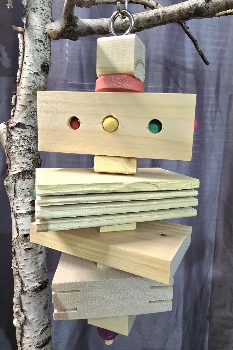 Puede incluir: Un juguete colgante para p&aacute;jaros hecho de madera natural, con una variedad de formas y colores. El juguete incluye un cubo de madera, un disco rosa y un bloque rectangular con tres agujeros de colores. Varias plataformas de madera est&aacute;n apiladas debajo.