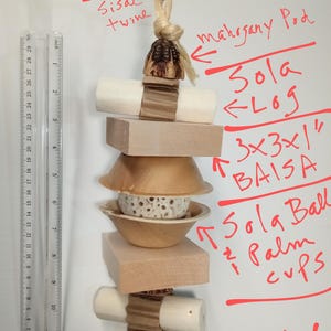 Pu&ograve; includere: Un giocattolo per uccelli fatto di materiali naturali, tra cui un tronco di sola, una palla di sola, tazze di palma e un blocco baisa di 7,6 x 7,6 x 2,5 cm. Il giocattolo &egrave; sospeso da un ramo con un gancio in acciaio inossidabile e spago di sisal. Il testo "Freddi's FAVE" &egrave; scritto sullo sfondo bianco.