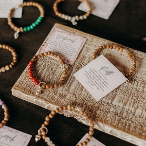 Könnte beinhalten: Sortiment aus Holz-Armbändern mit verschiedenen bunten Perlen und Charms. Jedes Armband wird mit einer kleinen Karte mit einem Wort und einer Beschreibung geliefert. Die Wörter umfassen "Glaube", "Hoffnung", "Liebe", "Freude", "Frieden" und "Stärke".